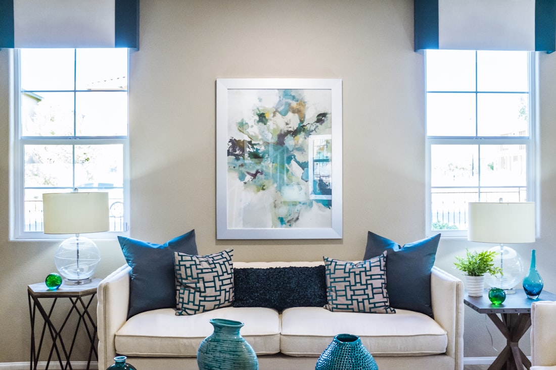 Cream sofa with blue cushions between two windows, under an abstract painting