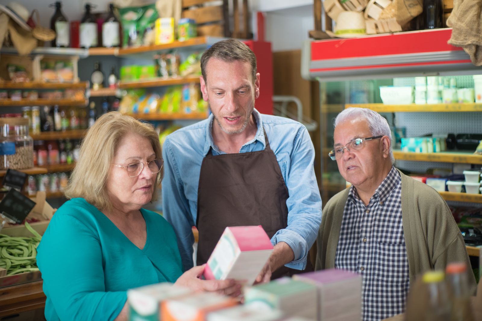 A shopkeeper in a brown apron shows an older couple a boxed product in a small grocery store