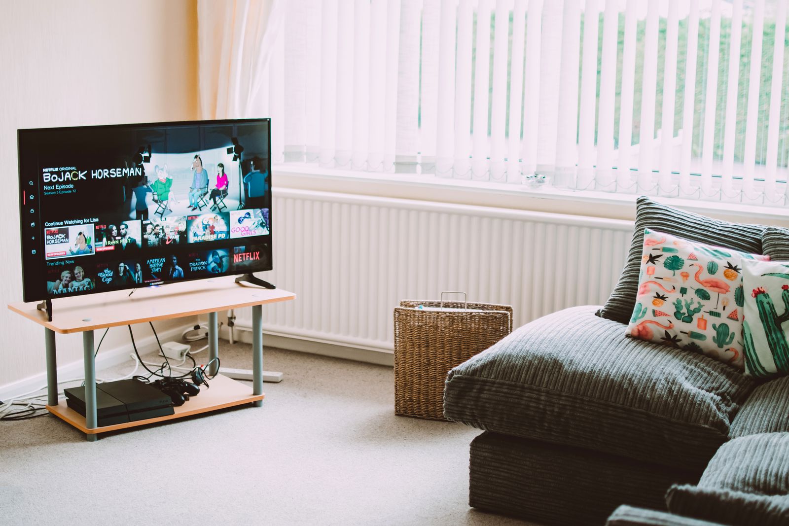 A living-room TV showing a streaming app menu on a low stand, a game console on the shelf and a grey sofa beside it