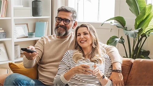 A couple smiling on a sofa, one holding a remote and the other a mug