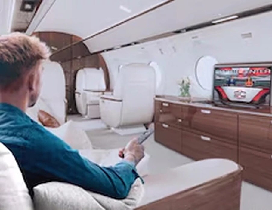 A man in a private-jet cabin pointing a remote at a TV that shows a sports broadcast